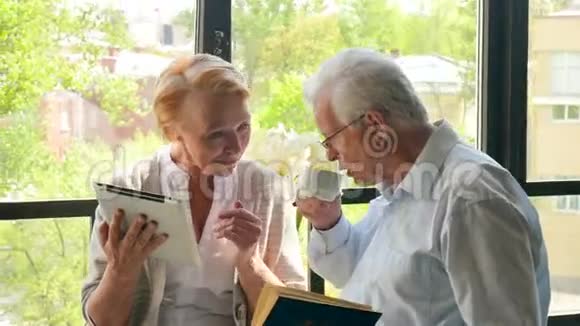 快乐可爱的退休夫妇站在对面拿着平板电脑和书他们微笑视频的预览图