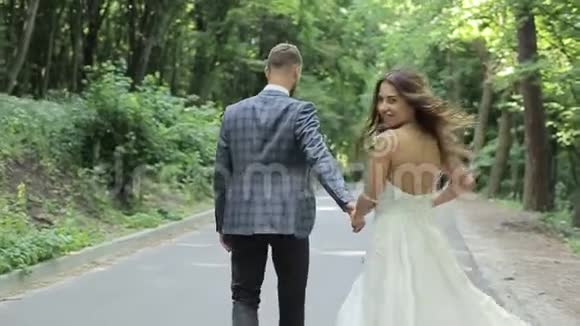 一对漂亮的已婚夫妇走在公园里牵着他们的手结婚礼服背景上的绿树视频的预览图