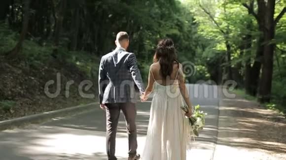一对漂亮的已婚夫妇走在公园里牵着他们的手结婚礼服背景上的绿树视频的预览图
