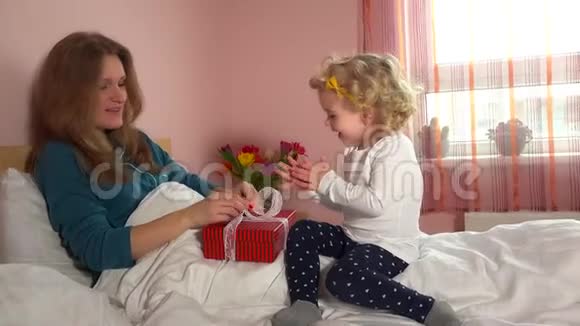快乐妈妈带着可爱的小女儿拆开礼物盒坐在床上视频的预览图