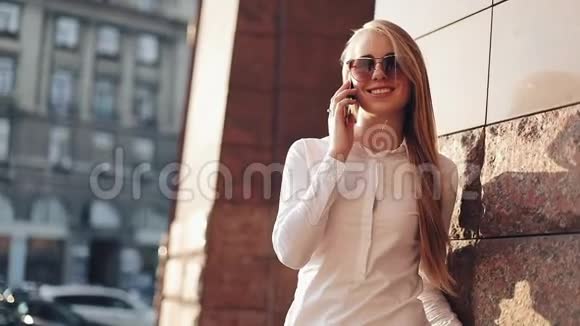 年轻的女商人站在阳光明媚的城市街道上的办公楼附近用手机交谈视频的预览图