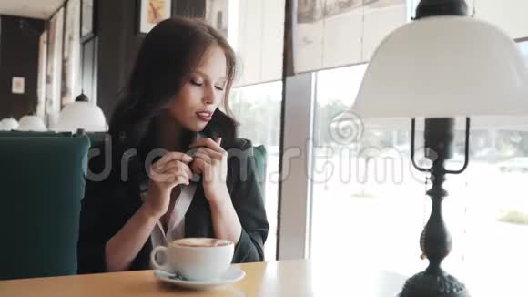 一位美丽的年轻女士在咖啡馆的窗户附近喝着泡沫咖啡的卡布奇诺咖啡视频的预览图