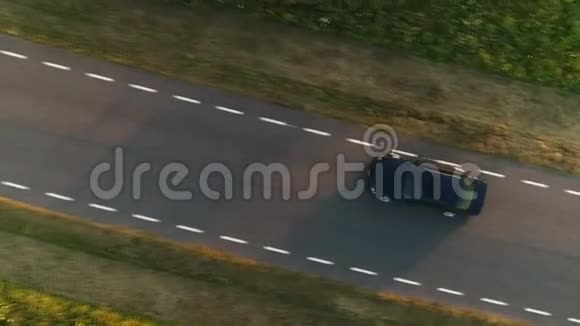 空中拍摄从上面驾驶黑色汽车在一个田野沿着乡村道路在夏天日落视频的预览图
