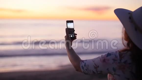 戴帽子的年轻女游客在海洋海滩日落时拍照视频的预览图