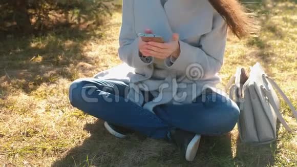 年轻漂亮的黑发女人坐在春天公园的草地上用手机输入一条信息特写双手视频的预览图