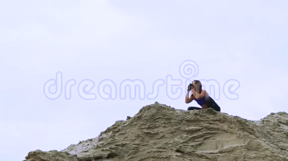 年轻的运动的女人穿着蓝色上衣和条纹腿裤做运动蹲在采石场的沙质顶部健身视频的预览图