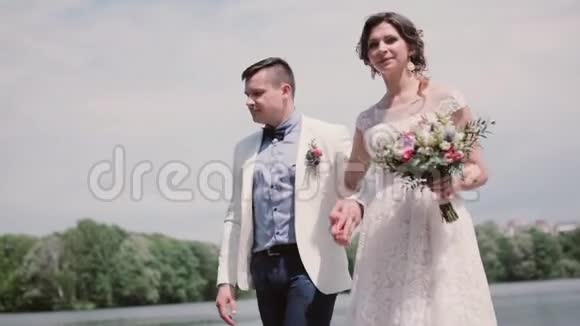 夏天美丽的新娘和英俊的新郎在公园里散步白色婚纱和礼服发型斯坦迪卡姆视频的预览图