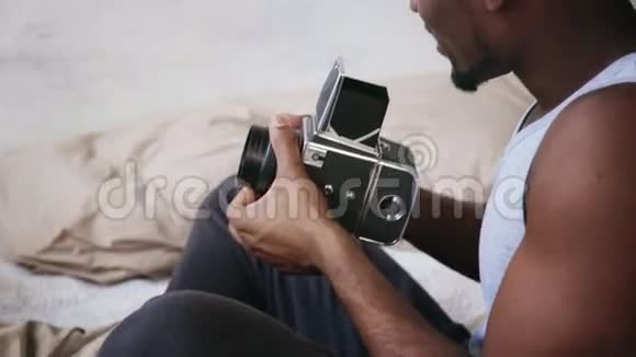 穿着睡衣的非洲年轻人坐在床上抱着旧的光镜有人在动影相机上说照片视频的预览图