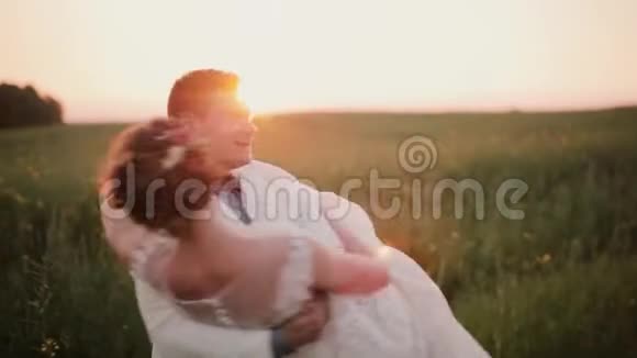 英俊的新郎把他美丽的新娘抱在怀里旋转着她笑着吻他自然日落视频的预览图
