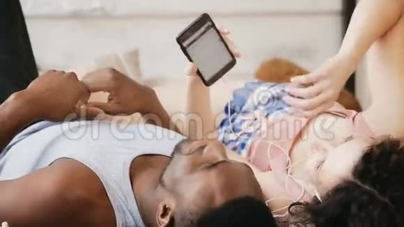 多种族夫妇躺在床上跳舞听音乐穿睡衣的男人和女人使用智能手机和耳机视频的预览图