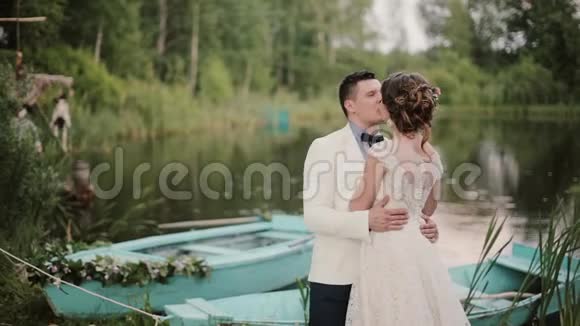 美丽的恋人在一个美丽的地方亲吻一个池塘池塘上有蓝色的船新娘和新郎享受他们的婚礼视频的预览图