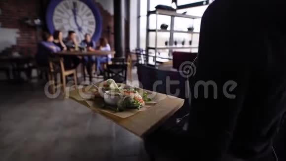 一个女服务员端上一张木桌桌子后面坐着一群男人和女人一个盘子里放着蔬菜小吃和酸味视频的预览图