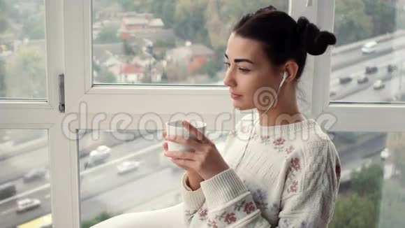 戴着耳机坐在窗边听音乐喝咖啡的迷人年轻女子视频的预览图