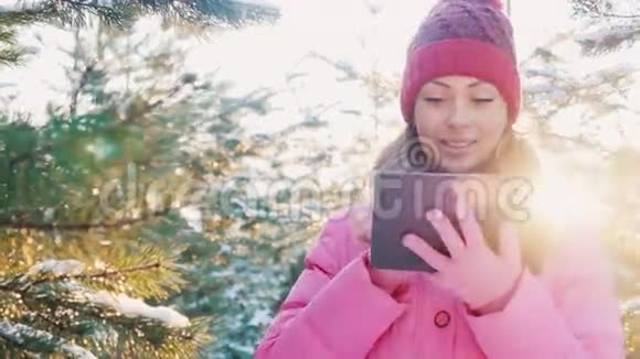 穿着粉红色冬衣的年轻女子喜欢平板电脑她站在冬天的森林里阳光照耀着视频的预览图