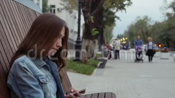 一个严肃的女孩坐在长凳上穿着牛仔裤用手机发短信视频的预览图