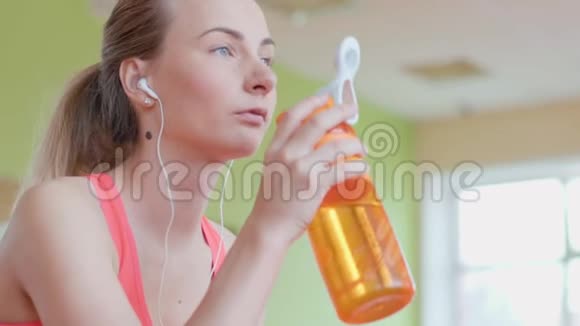 穿着运动服戴着耳机的漂亮女孩在运动后休息时一边听音乐一边喝水视频的预览图