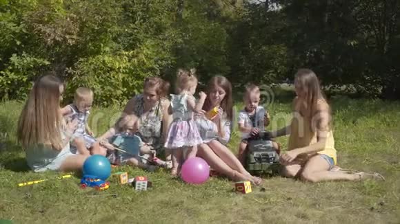 一群快乐的孩子在夏季公园户外玩耍母亲们照看坐在草地上的孩子视频的预览图