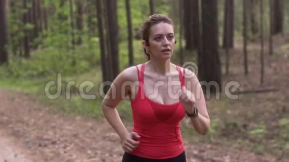 年轻迷人的女人在夏天的森林里慢跑带无线耳机的女孩和带心率监视器的手表视频的预览图