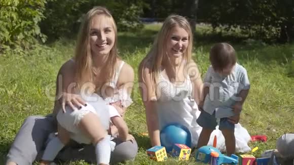 一群快乐的孩子在夏季公园户外玩耍母亲们照看坐在草地上的孩子视频的预览图