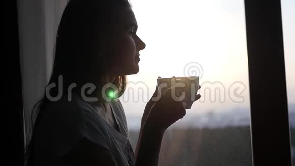 坐在窗边的一个年轻迷人的女孩欣赏着夕阳捧着一杯热茶1920x1080慢动作视频的预览图