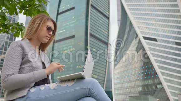 一个金发女孩在市中心和高楼的背景下使用白色笔记本电脑戴太阳镜的女孩视频的预览图