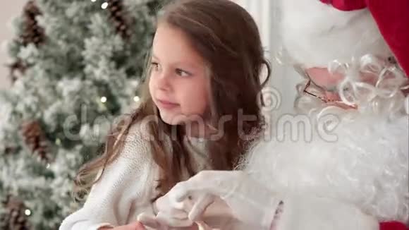 圣诞老人在礼物上系上丝带蝴蝶结送给一个坐在家中圣诞树旁的小女孩视频的预览图