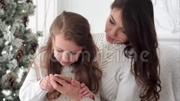 年轻的母亲教女儿如何在圣诞节装饰的房间里使用智能手机视频的预览图