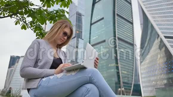 一个金发女孩在市中心和高楼的背景下使用白色笔记本电脑戴太阳镜的女孩视频的预览图