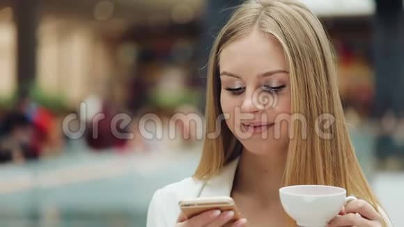 迷人的年轻女子一手拿着智能手机一手拿着咖啡坐在咖啡馆里视频的预览图