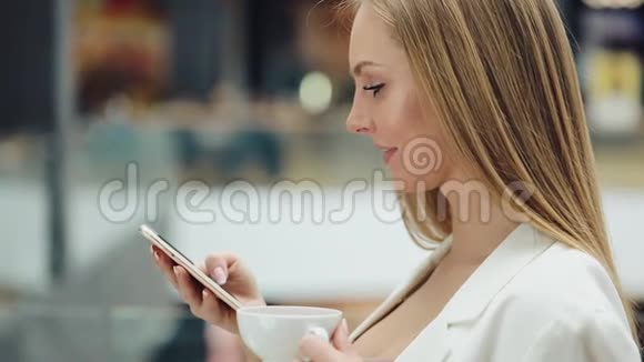 迷人的年轻女子一手拿着智能手机一手拿着咖啡坐在咖啡馆里视频的预览图
