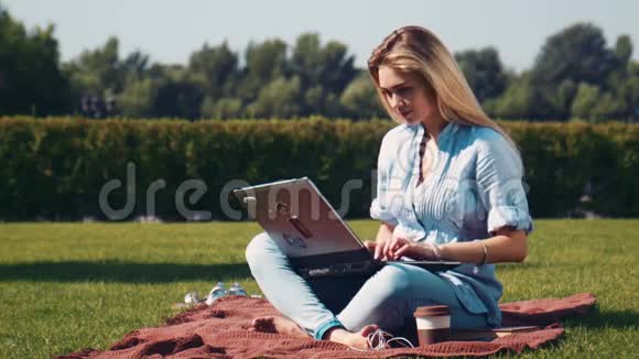 带着笔记本电脑在公园的毯子上微笑的漂亮女孩视频的预览图