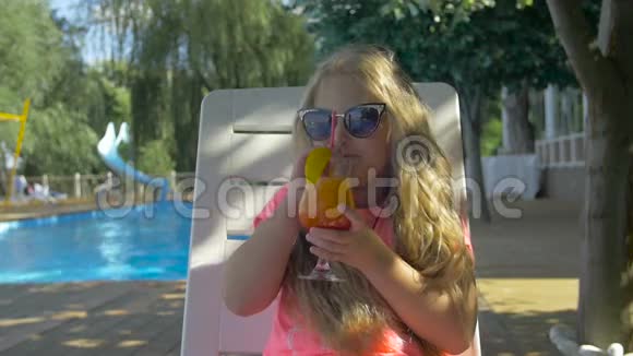 小漂亮的女孩坐在游泳池附近的椅子上喝着新鲜的橙汁可爱的孩子享受她的夏天视频的预览图