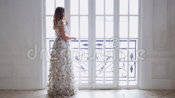 金发女孩穿着一件明亮的长裙子穿着一扇大窗户视频的预览图