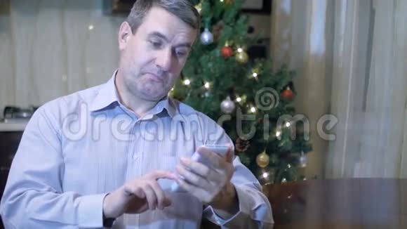 一个英俊的中年男子手里拿着一部手机坐在家里的餐桌上背景是圣诞树视频的预览图