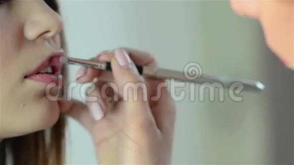 化妆师在一位年轻漂亮女人的嘴唇上用刷子涂红色口红特写镜头视频的预览图