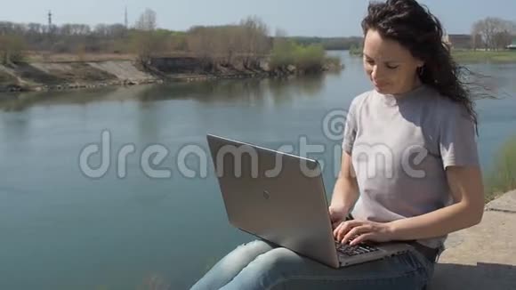 带着笔记本电脑在河边的年轻女子女孩坐在河岸上拿着笔记本电脑一个春天阳光明媚的日子视频的预览图
