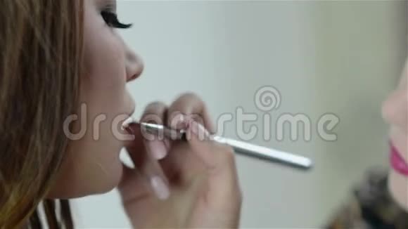 化妆师在一位年轻漂亮女人的嘴唇上用刷子涂红色口红特写镜头视频的预览图