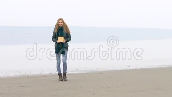 年轻漂亮的女孩走在沙滩上拍照带着盒子微笑视频的预览图