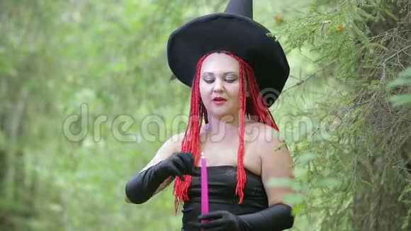 带着红色头发和尖尖帽子的迷人年轻女巫在森林里用蜡烛召唤视频的预览图