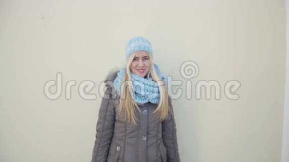 年轻女子穿着浅蓝色的冬帽和围巾靠在墙上微笑着拥抱着相机视频的预览图