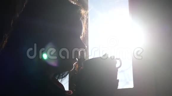 美丽的女孩享受着一杯美味的热茶站在窗边晒太阳从杯子里喝慢动作视频的预览图