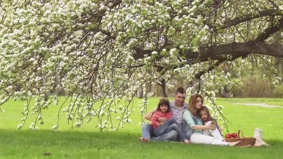爸爸妈妈和孩子们坐在一棵盛开的苹果树下的草地上幸福的家庭在大自然中休息二视频的预览图