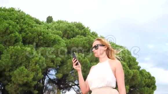 戴着太阳镜的优雅的金发女人对着天空和树叶打电话从下面看4k慢视频的预览图