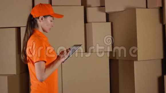 女性仓库工作人员穿着橙色制服计数箱并使用她的平板电脑与棕色纸箱背面4K视频视频的预览图