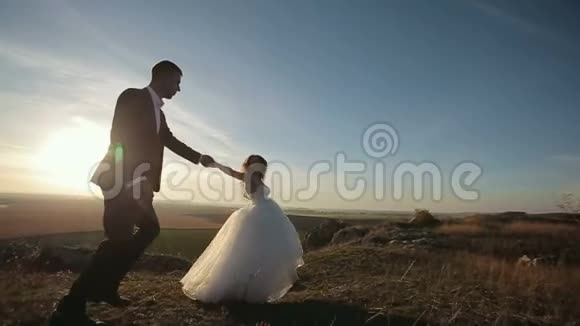 新娘和新郎牵着手在夕阳下沿着山岩奔跑视频的预览图