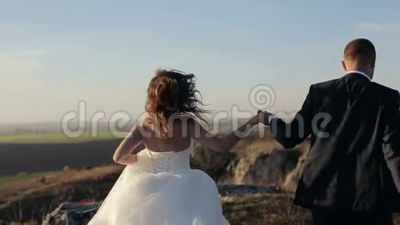 新娘和新郎牵着手在夕阳下沿着山岩奔跑视频的预览图