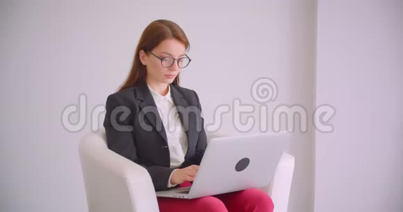 戴着眼镜的年轻白种人女商人的特写肖像她用笔记本电脑看着坐在扶手椅上的照相机视频的预览图