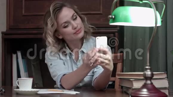 年轻的女人坐在桌旁拿着书拿着手机玩着滑稽的自拍视频的预览图