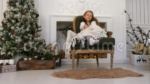 微笑的女孩裹着毯子坐在舒适的椅子上等待圣诞老人视频的预览图