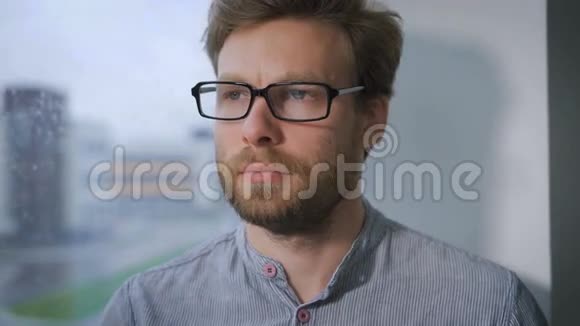 戴眼镜的年轻人脸上带着沉思的表情坐在窗台上视频的预览图